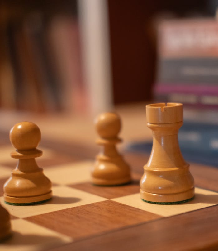 services-02 Detailed view of chess pieces on a wooden chessboard with books in the background.